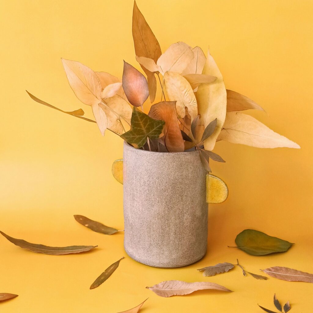 Autumn still life with dried leaves arranged in a ceramic vase on a warm yellow surface. Cozy seasonal image for wallpapers, fall branding, posters, web design, social media visuals, and editorial use.